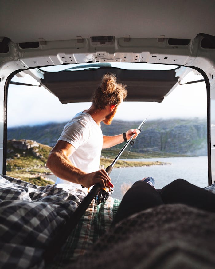 home-hero-2 Man enjoys fishing from a van with a scenic lake view, embracing outdoor adventure and travel.