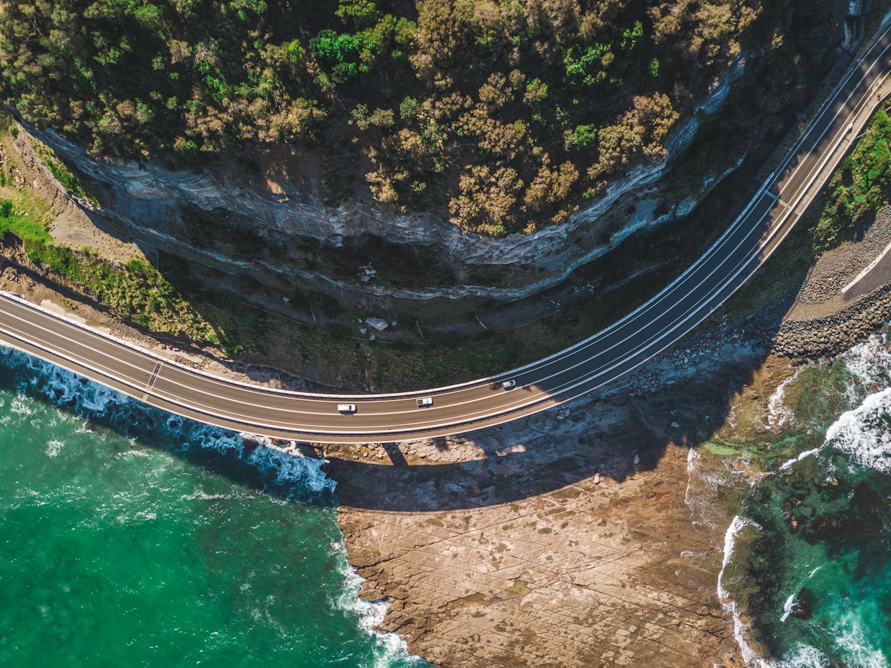 about-us-01 A breathtaking aerial view of a winding coastal road in Clifton, NSW, Australia with turquoise sea and rugged cliffs.