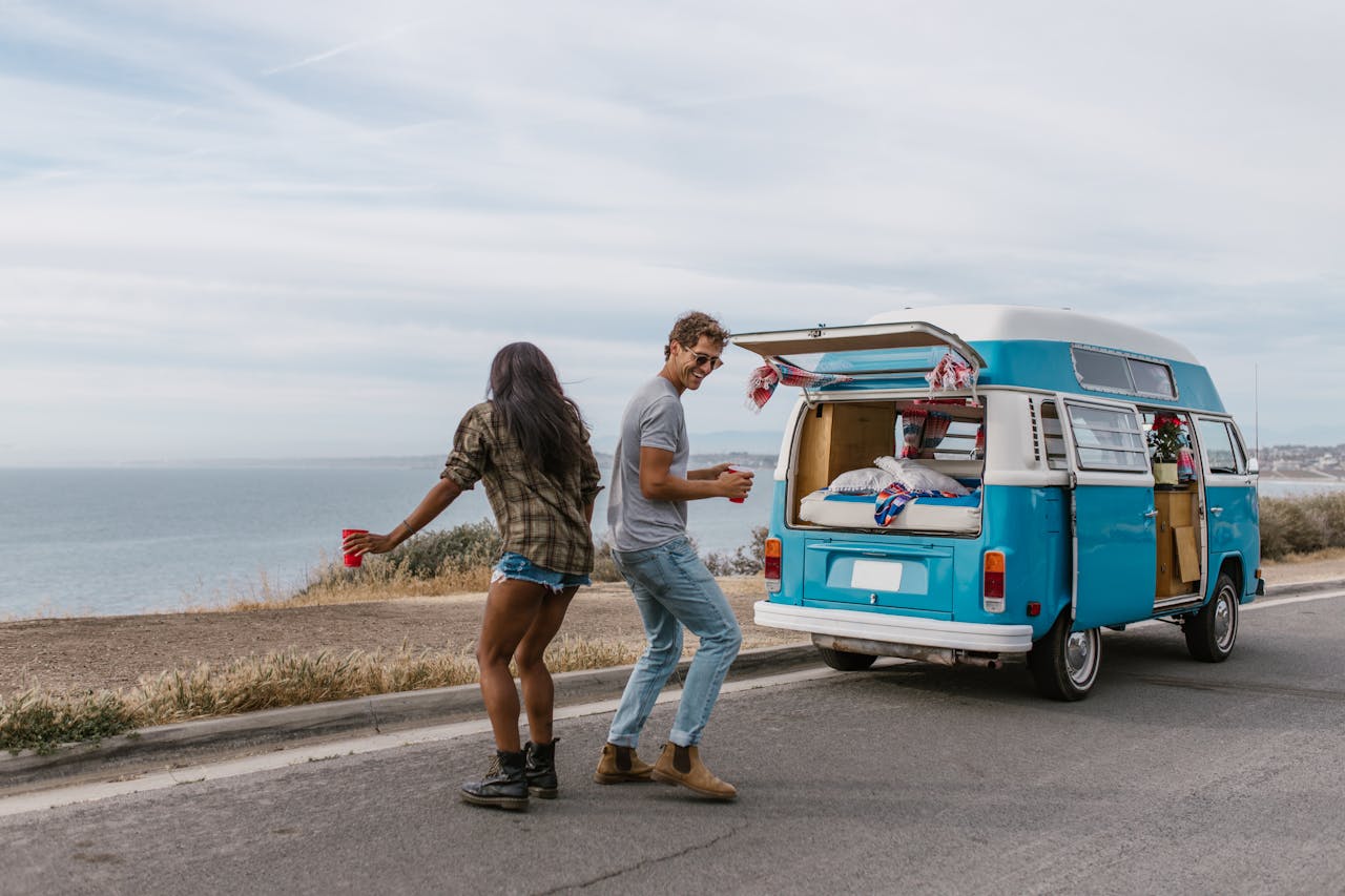 home-hero-1 Happy couple dancing by their blue campervan on a scenic coastal road trip.