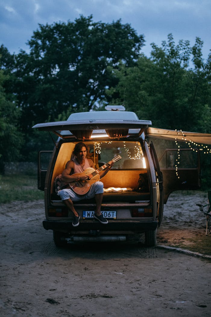 services-04 A young man sits in a van playing guitar surrounded by nature at dusk.